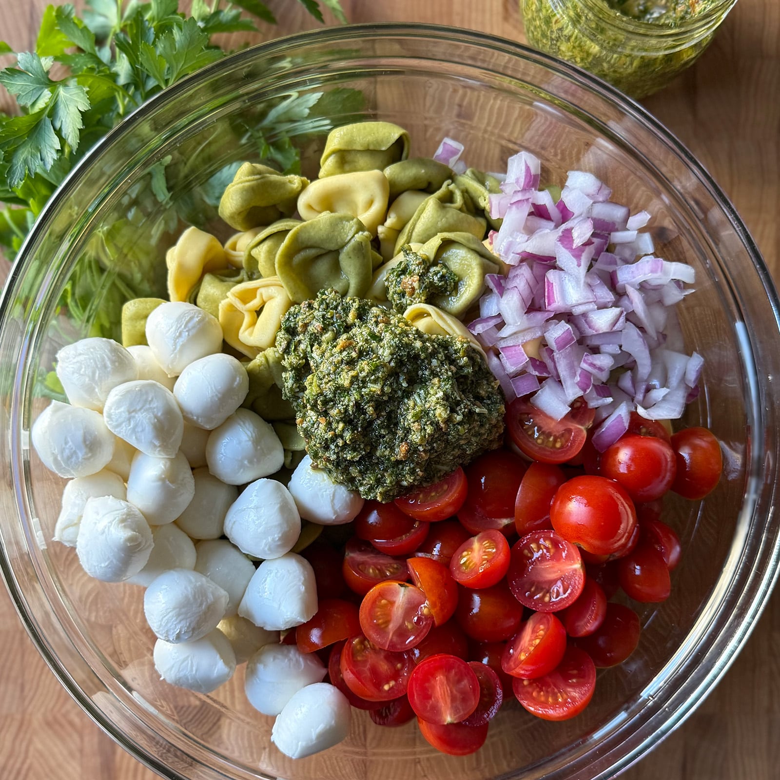 A large glass bowl with cooked tortellini, halved cherry tomatoes, mini mozzarella balls, diced red onion, and a scoop of mint pesto before tossing