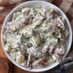 Overhead view of red potato salad in a white bowl on a wooden cutting board, garnished with fresh dill and chives.