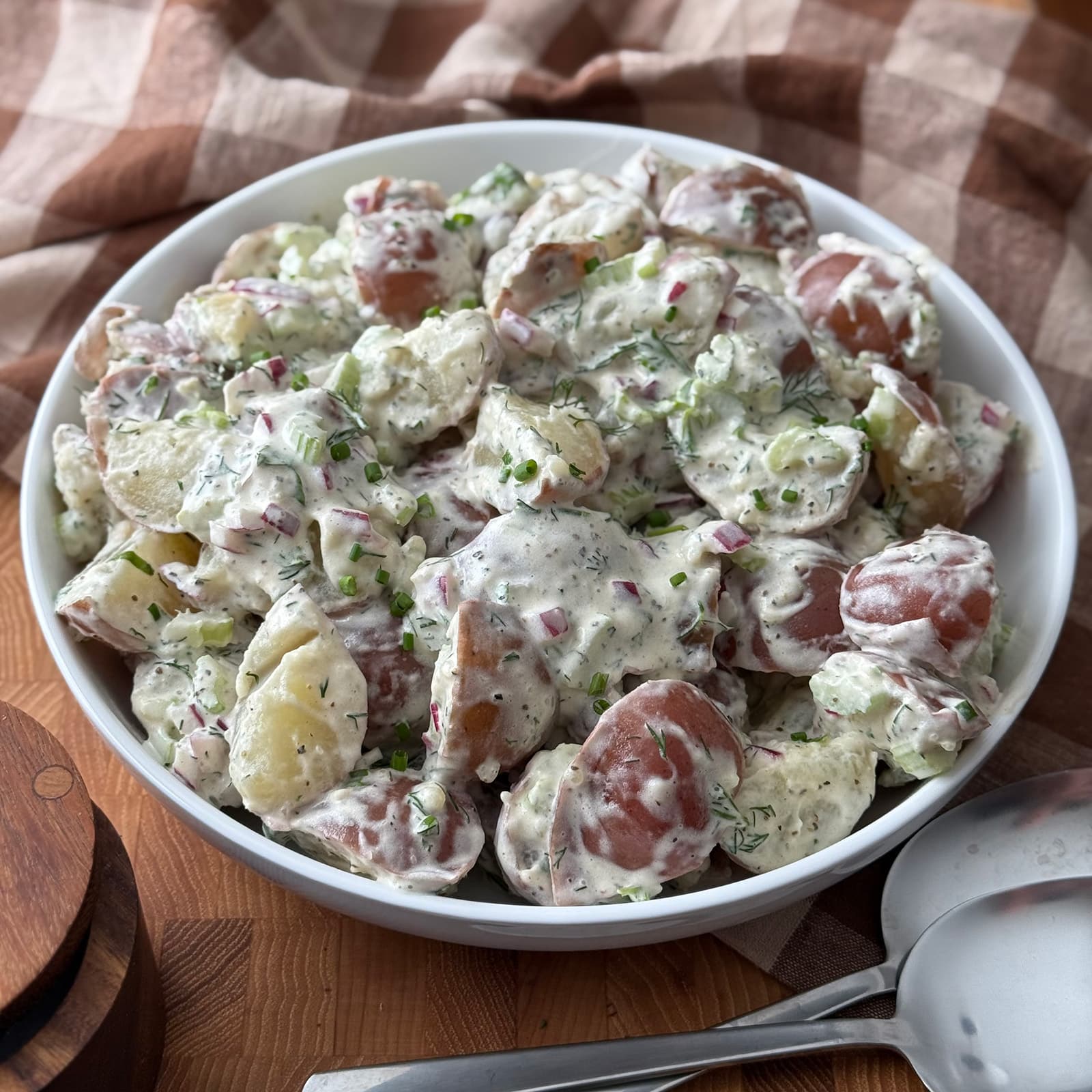 Red skinned potato salad in a white bowl on a wooden cutting board with a serving spoon, surrounded by a plaid linen napkin.