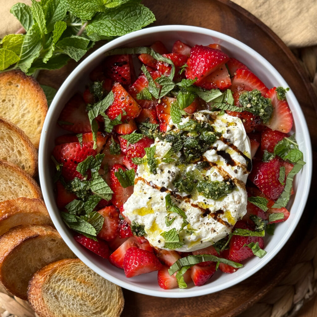 Overhead view of strawberry burrata salad in a white bowl on a wooden serving board, with fresh strawberries, creamy burrata, torn mint, pesto drizzle, and balsamic glaze, surrounded by toasted baguette slices and a sprig of fresh mint