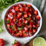 Overhead view of fresh strawberry salsa in a white bowl with diced strawberries, jalapeño, red onion, and cilantro, surrounded by whole strawberries and a halved lime on a linen cloth