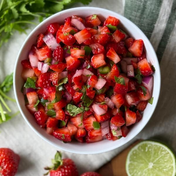 Overhead view of fresh strawberry salsa in a white bowl with diced strawberries, jalape&ntilde;o, red onion, and cilantro, surrounded by whole strawberries and a halved lime on a linen cloth