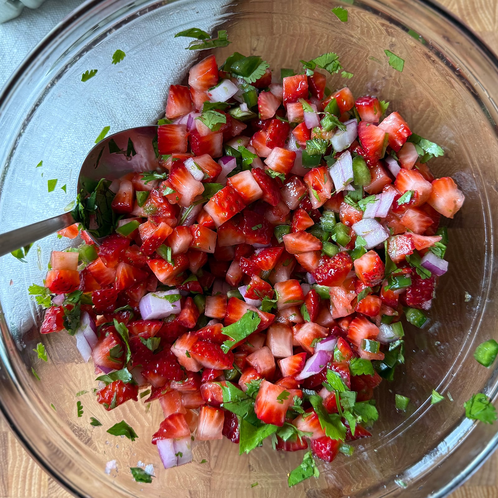 Strawberry salsa being stirred with a spoon in a large clear glass mixing bowl, showing diced strawberries, red onion, jalape&ntilde;o, and cilantro