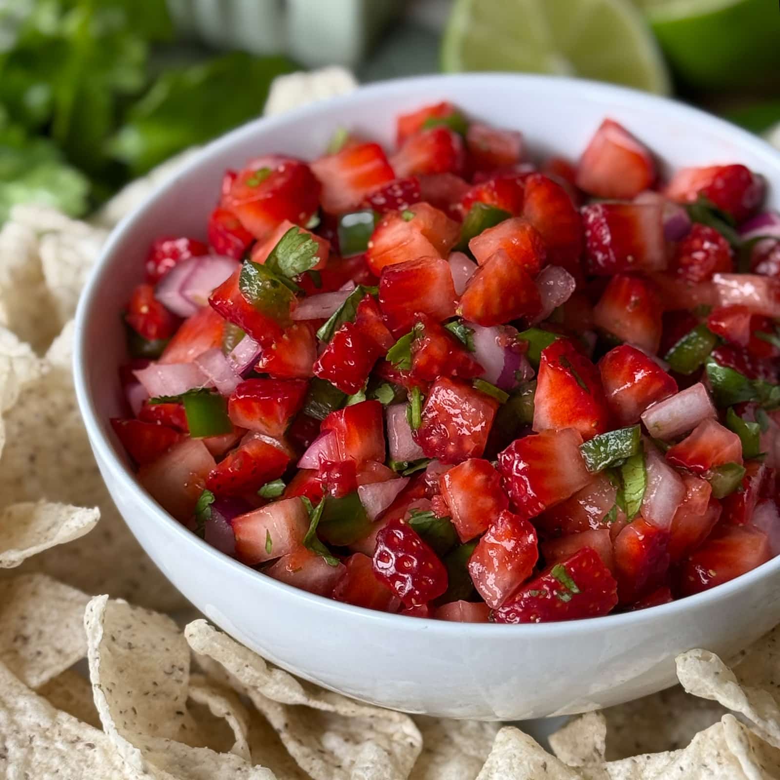 Strawberry salsa served in a white bowl surrounded by tortilla chips with fresh cilantro and lime visible in the background