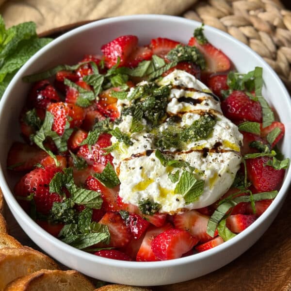 Angled close-up of strawberry burrata salad in a white bowl showing creamy burrata broken open and topped with dairy-free pesto, balsamic glaze, torn fresh mint, and cracked black pepper, with toasted baguette slices and a woven placemat visible in the background