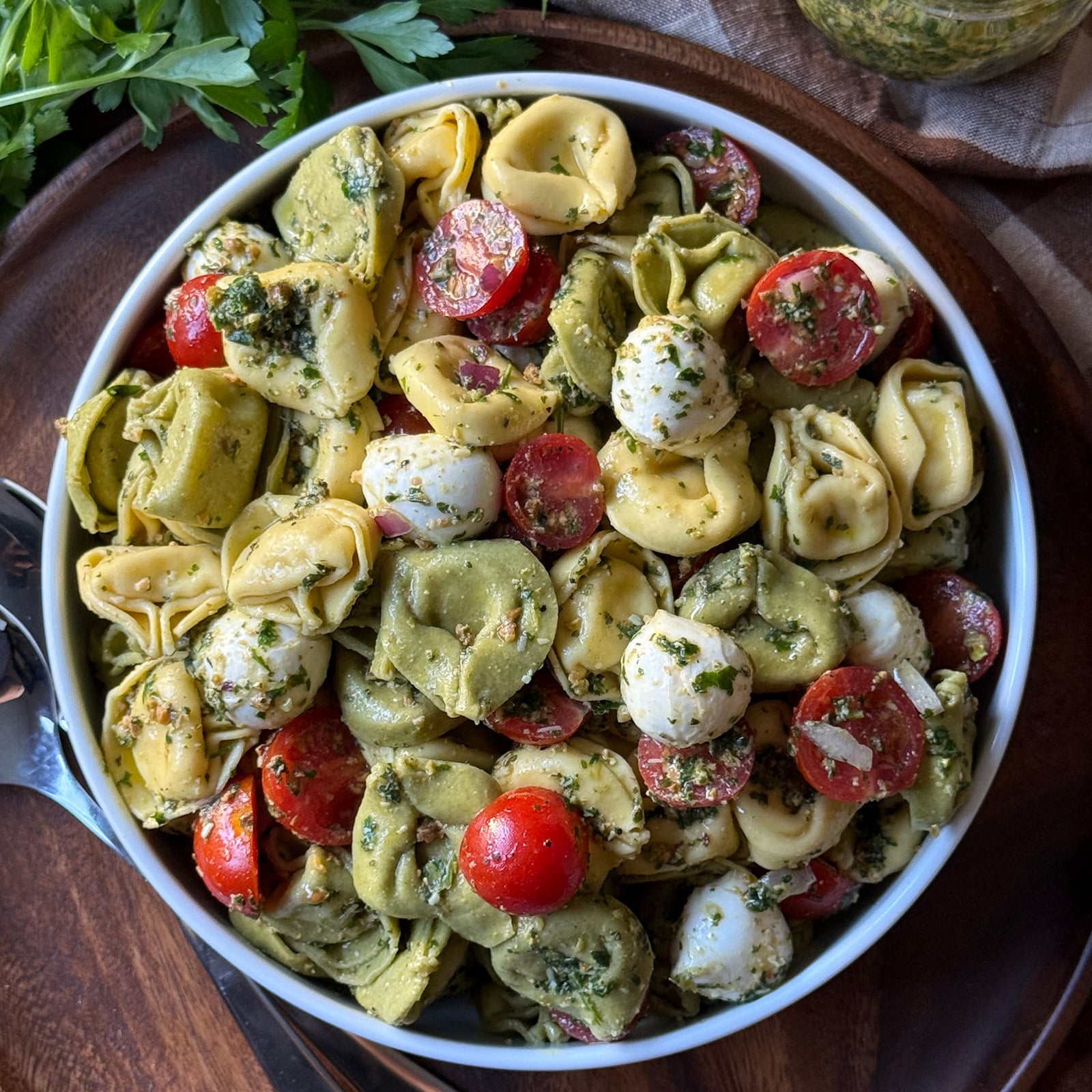 Overhead shot of a white bowl filled with Tortellini Pasta Salad with Pesto, showing tri-color tortellini, cherry tomatoes, mozzarella balls, and fresh parsley coated in mint pesto