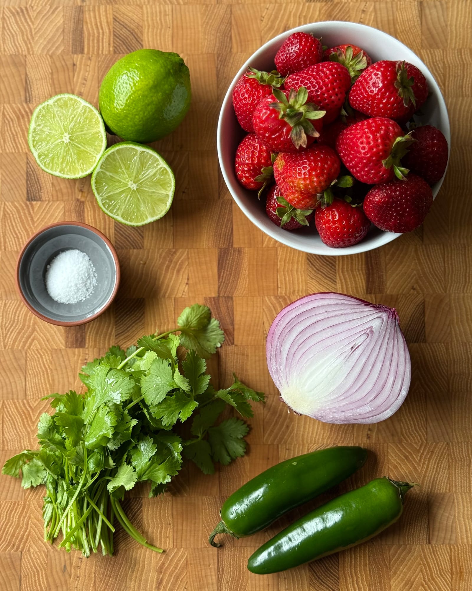 Flat lay of whole strawberry salsa ingredients on a butcher block cutting board including fresh strawberries in a white bowl, two jalape&ntilde;os, a halved red onion, cilantro, limes, and a small dish of kosher salt