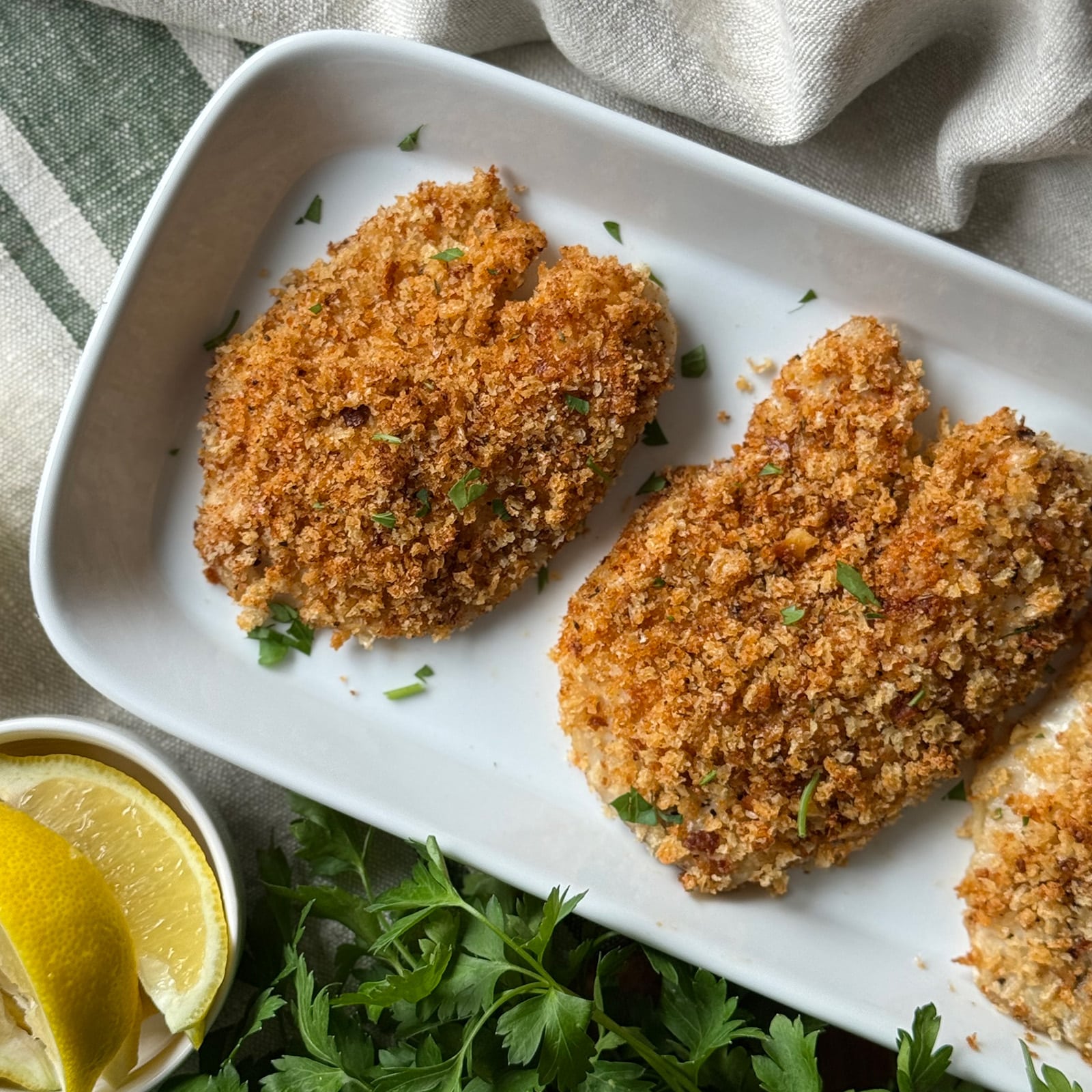 Three crispy Cajun breaded tilapia fillets on a white rectangular platter garnished with chopped fresh herbs and served alongside lemon wedges