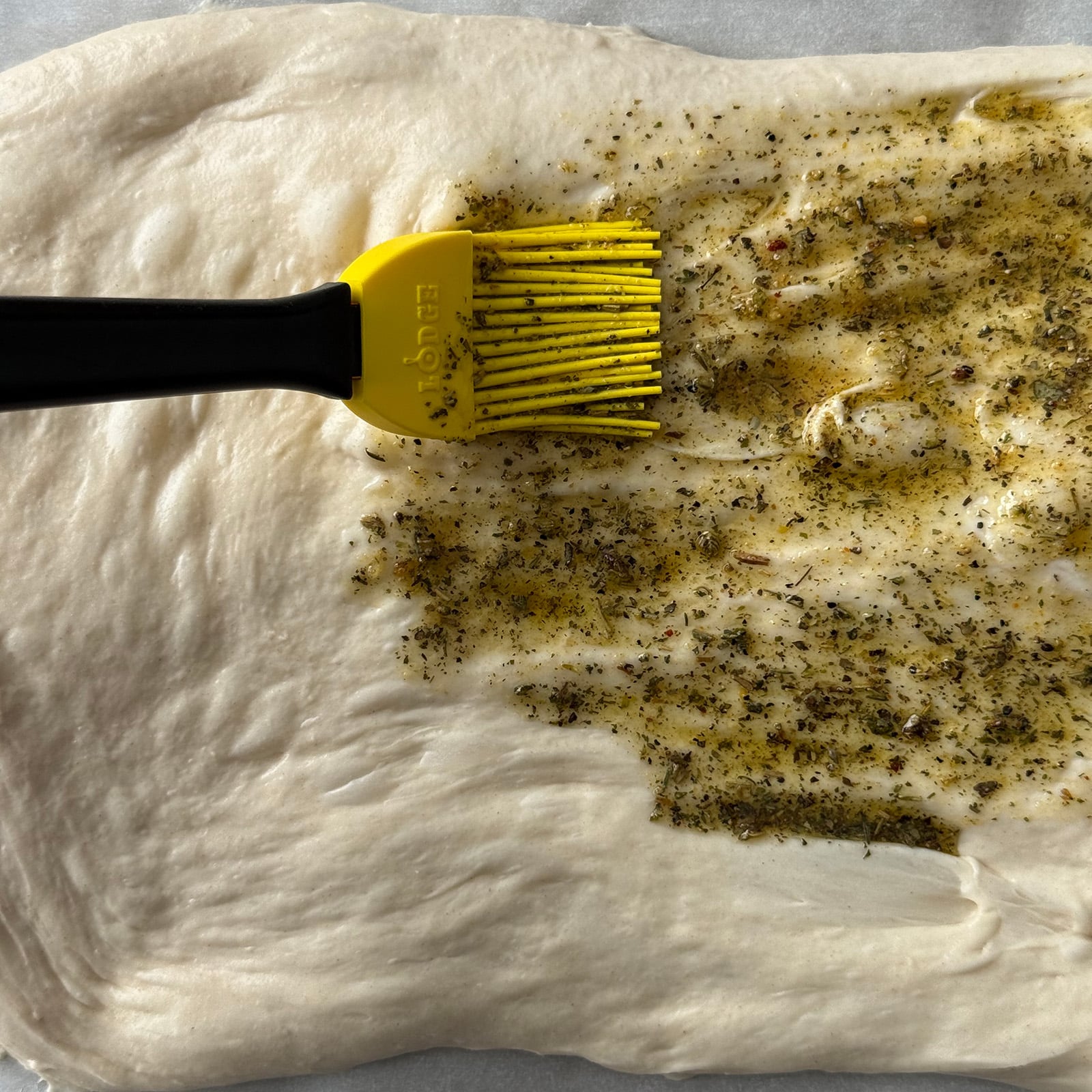 A silicone pastry brush spreading an Italian seasoning and olive oil mixture over stretched pizza dough on parchment paper.
