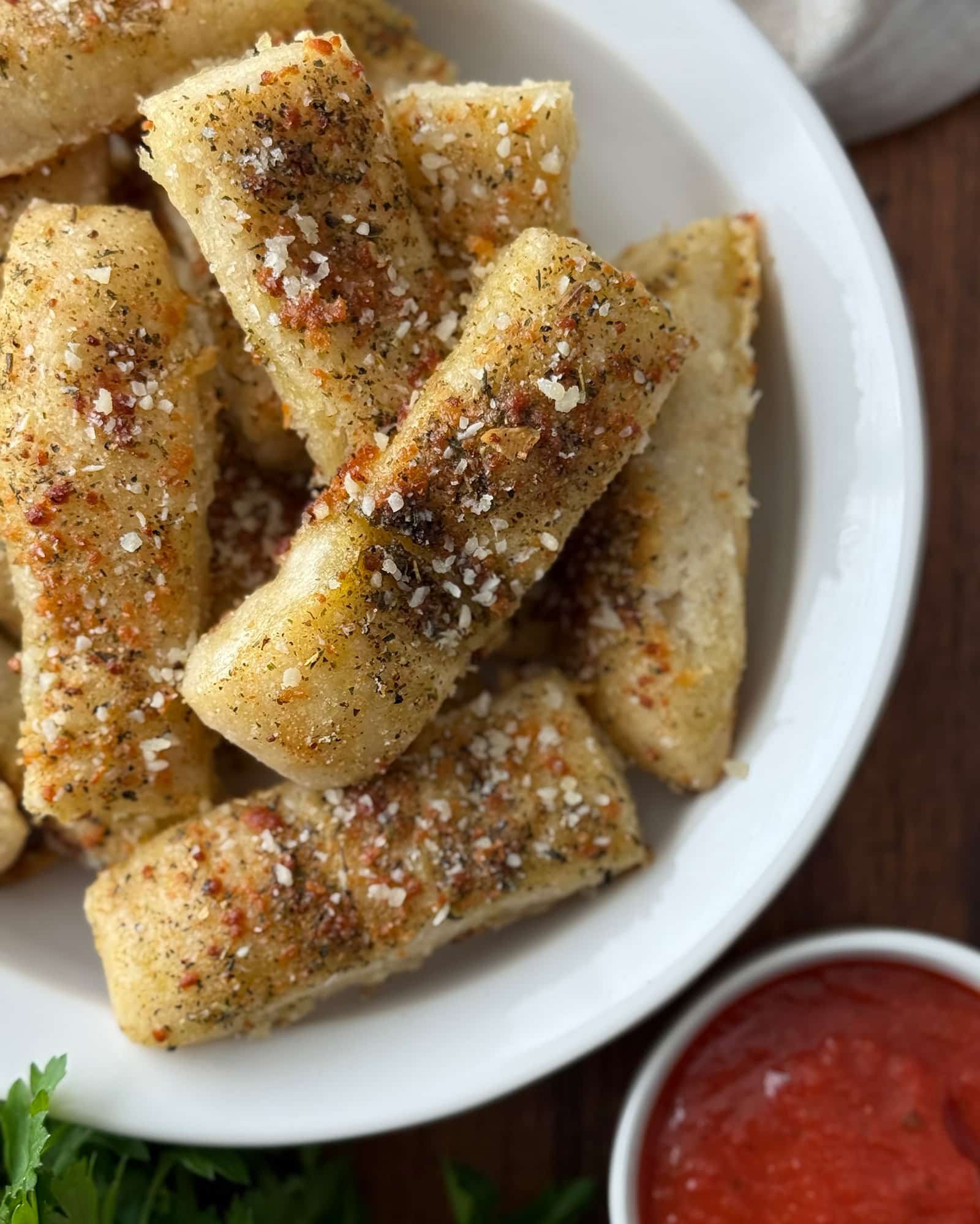 Garlic breadsticks from pizza dough piled in a white bowl, seasoned with Italian herbs and grated parmesan, with a small bowl of marinara sauce and fresh parsley alongside.