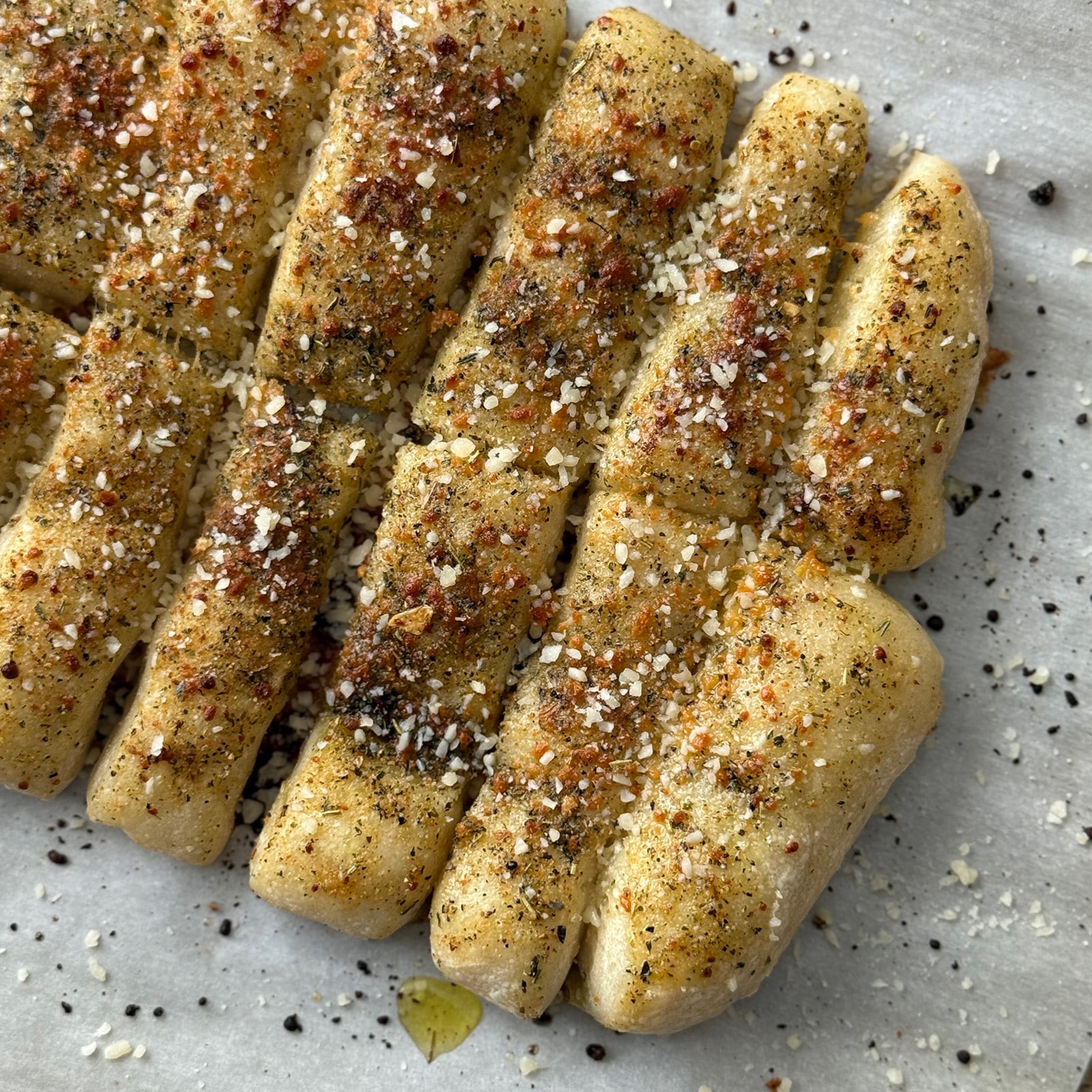 Freshly baked garlic breadsticks on parchment paper topped with grated parmesan cheese and Italian seasoning.