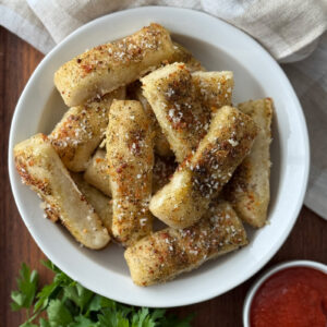 Garlic breadsticks from pizza dough piled in a white bowl on a wooden surface, seasoned with Italian herbs, granulated garlic and grated parmesan, served alongside a small bowl of marinara sauce