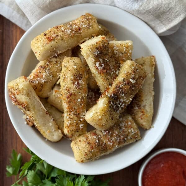 Garlic breadsticks from pizza dough piled in a white bowl on a wooden surface, seasoned with Italian herbs, granulated garlic and grated parmesan, served alongside a small bowl of marinara sauce