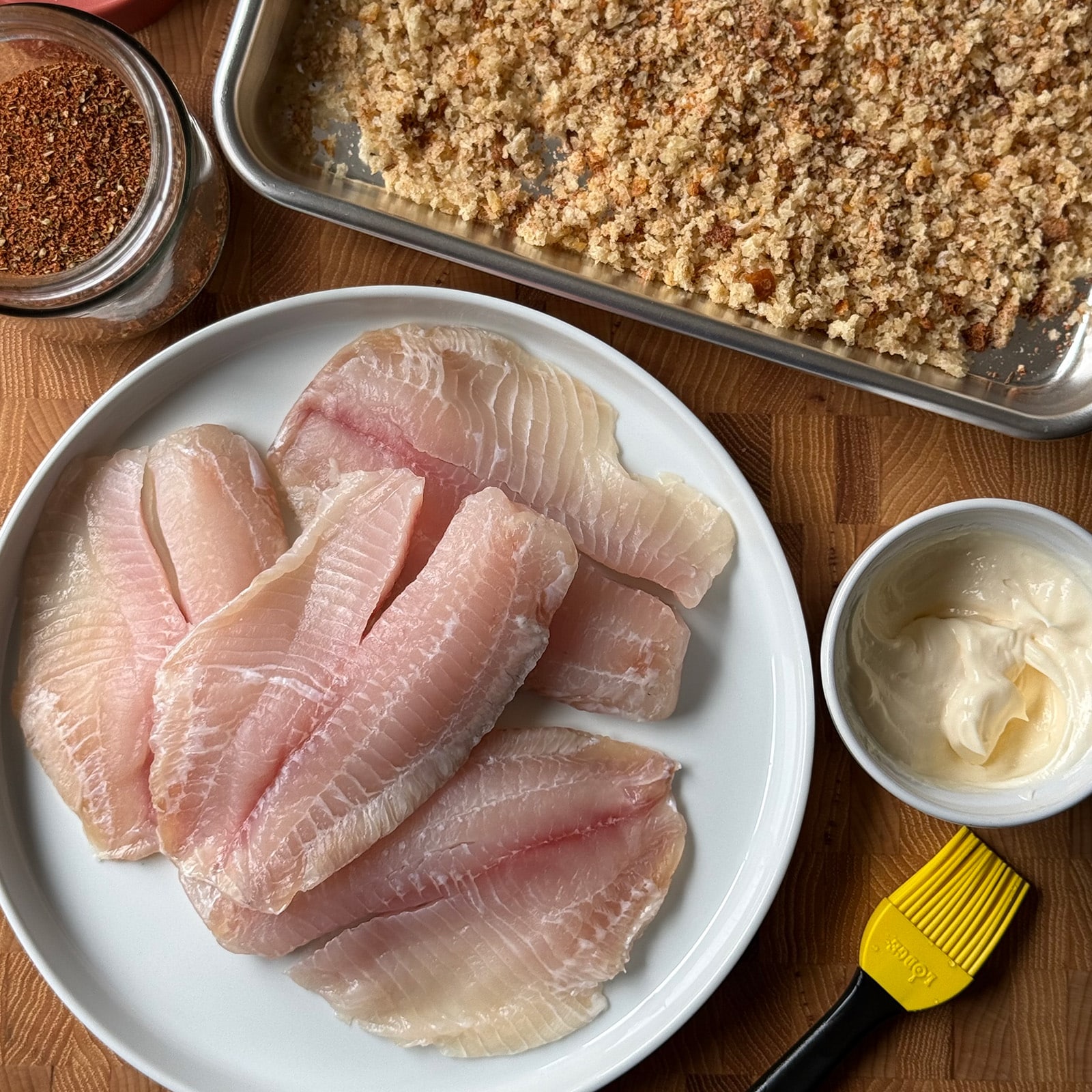 Ingredients for Cajun breaded tilapia including raw tilapia fillets, mayonnaise, Cajun spice blend, and seasoned panko breadcrumbs on a wood cutting board