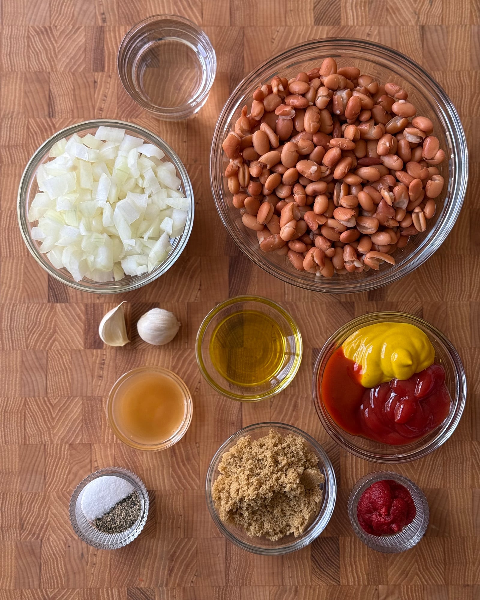 Overhead lay flat of ingredients for vegetarian baked beans, including pinto beans, diced onion, ketchup, mustard, brown sugar, tomato paste, garlic, olive oil, apple cider vinegar, salt, and pepper.