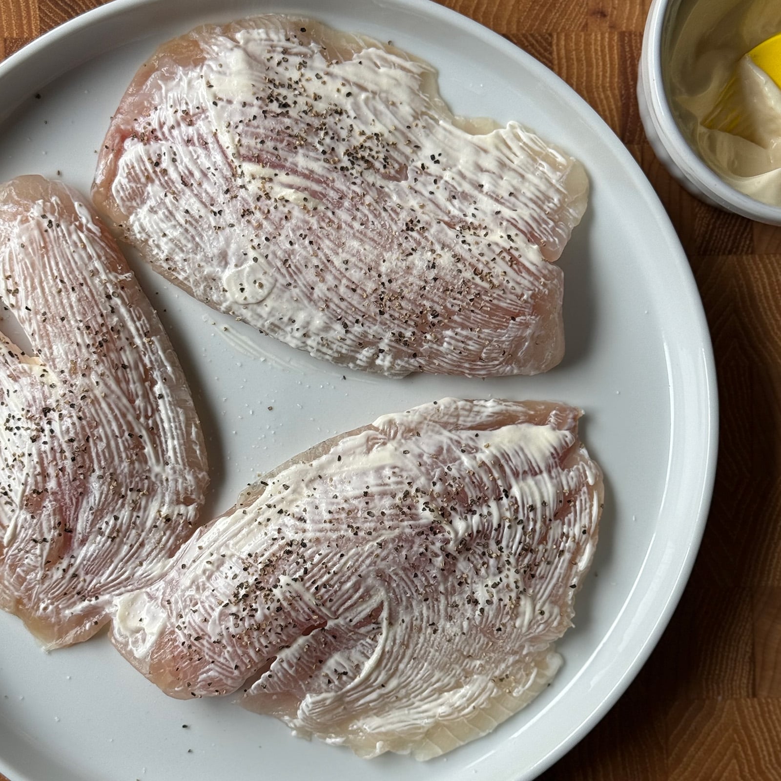 Three raw tilapia fillets brushed with mayonnaise and seasoned with black pepper on a white plate next to a small bowl of mayonnaise