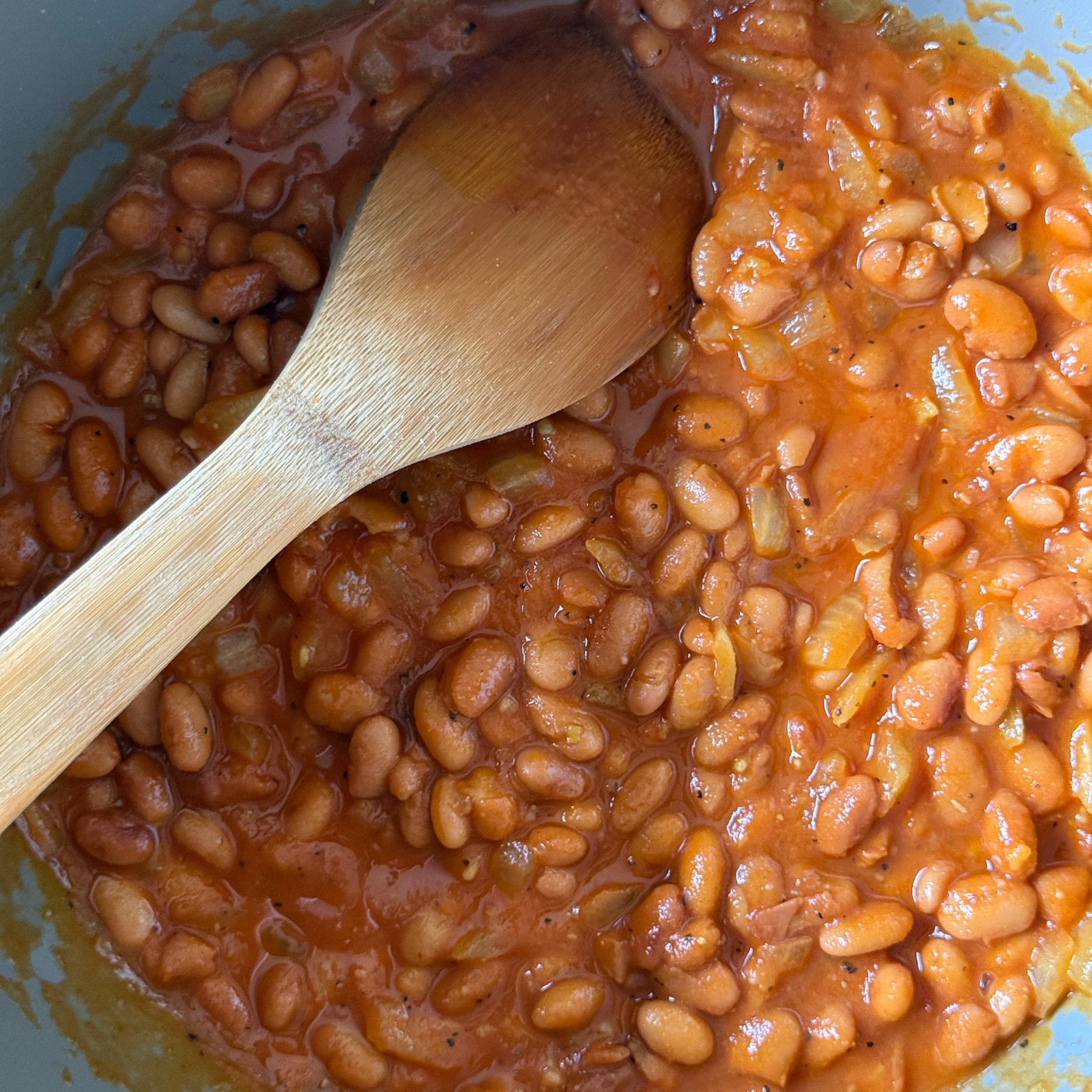 Vegetarian baked beans simmering in a pot with a wooden spoon, sauce thickened and beans glossy and tender.