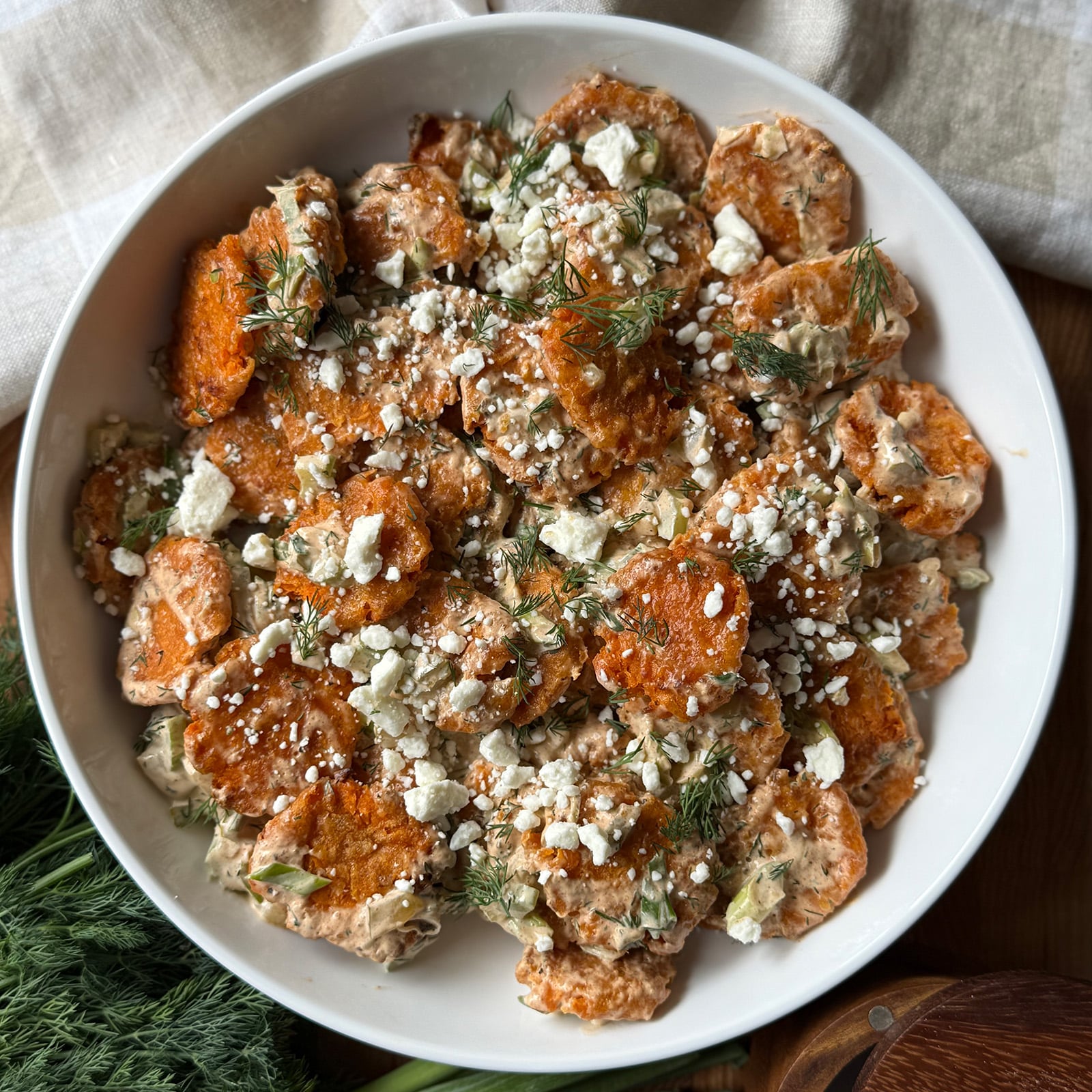 Overhead view of smashed sweet potato tater tot salad in a white bowl, topped with crumbled feta and fresh dill, with green onions and dill sprigs arranged alongside the bowl.