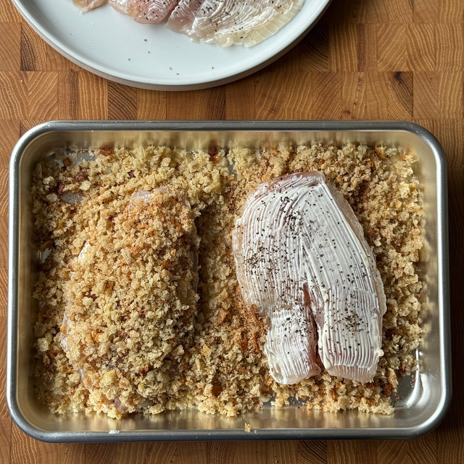 Two tilapia fillets in a rimmed baking sheet of seasoned Cajun panko breadcrumbs. One fully coated in the crust and one mayo-coated and ready to be pressed in.