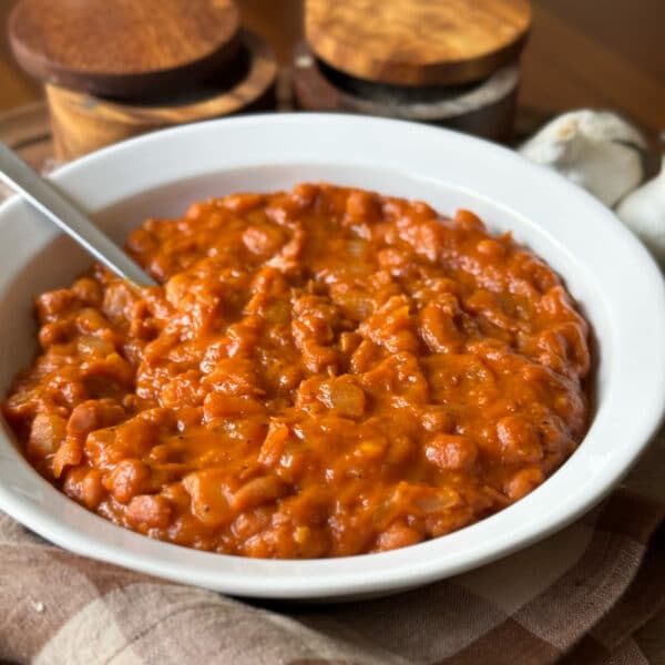 A bowl of vegetarian baked beans topped with a spoon, showing thick, glossy tomato-based sauce coating tender pinto beans, with garlic and wooden salt and pepper cellars in the background.