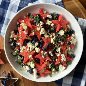 A white bowl filled with star-shaped watermelon, fresh blueberries, crumbled feta, and chopped basil and mint, with red and silver star-shaped cookie cutters on a blue plaid napkin alongside.