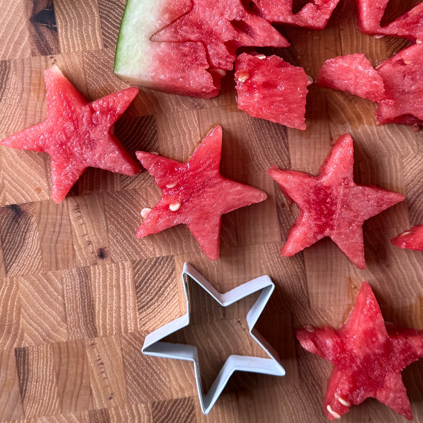 Star-shaped watermelon pieces on a wood cutting board next to a star-shaped cookie cutter, with watermelon scraps and a large watermelon slice in the background.