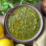 Overhead shot of finished chermoula sauce in a gray bowl with a gold spoon, surrounded by fresh parsley and lemons.