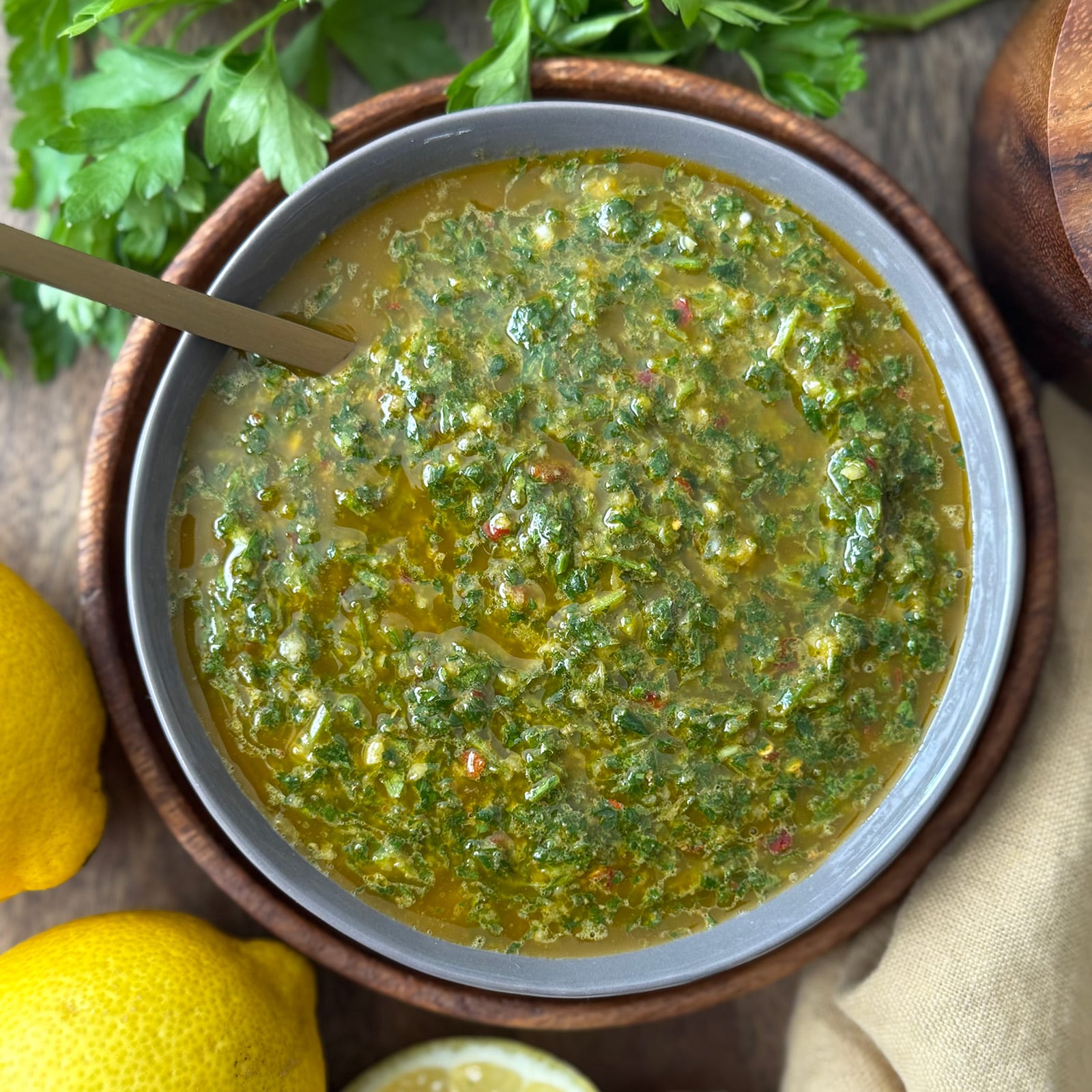 Overhead shot of finished chermoula sauce in a gray bowl with a gold spoon, surrounded by fresh parsley and lemons.
