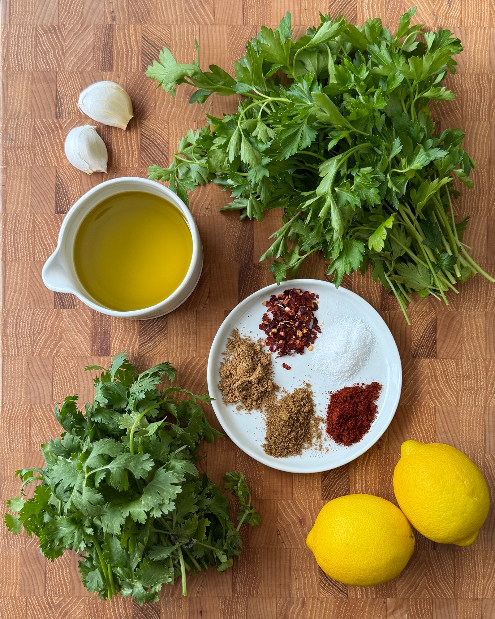 Overhead flat lay of chermoula ingredients on a wood cutting board, including fresh parsley, cilantro, garlic, two lemons, olive oil, and a plate of ground spices.