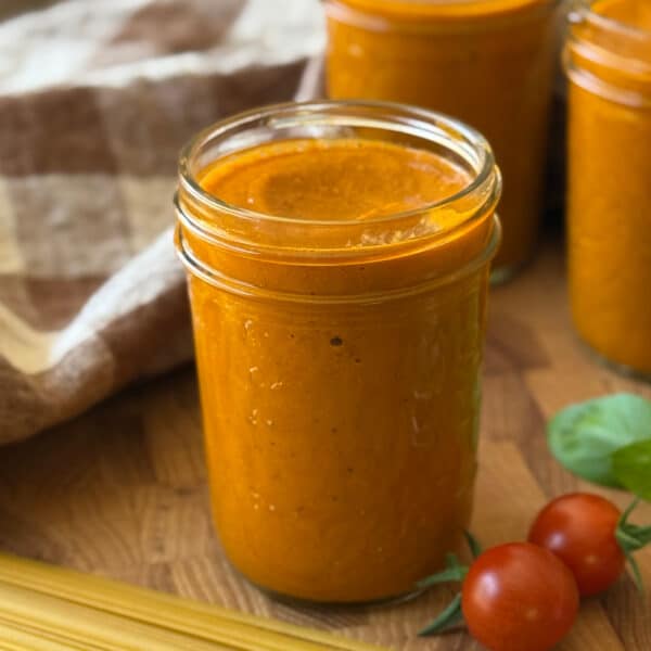 A glass jar filled with creamy Cherry Tomato Sauce sits on a wooden surface near fresh tomatoes, basil leaves, uncooked pasta, and a checkered cloth in the background.