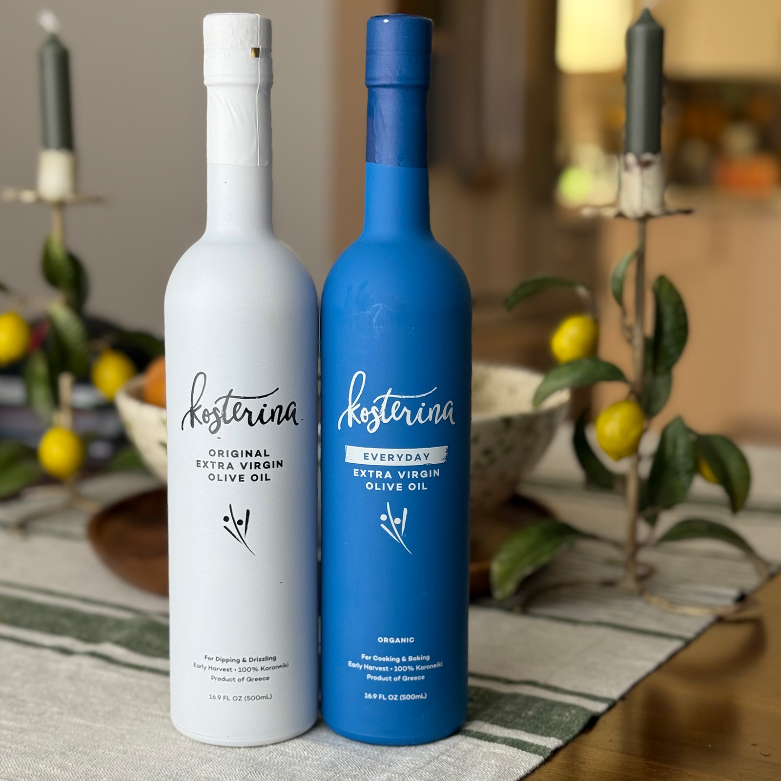 Two Kosterina olive oil bottles, one white and one blue, are side by side on a table with a bowl of fruit and two lit candles in the background. The bottles are labeled Original and Everyday, perfect for drizzling over pesto or roasted potatoes to elevate any meal.