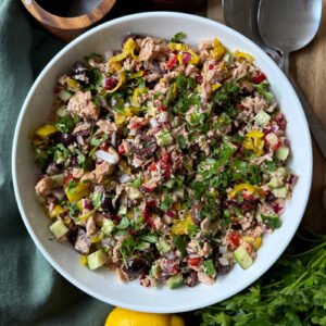 A white bowl filled with a colorful Mediterranean salad, including cucumber, olives, tuna, red onion, herbs, and pepperoncini. Fresh parsley, lemon, and serving utensils are visible nearby.