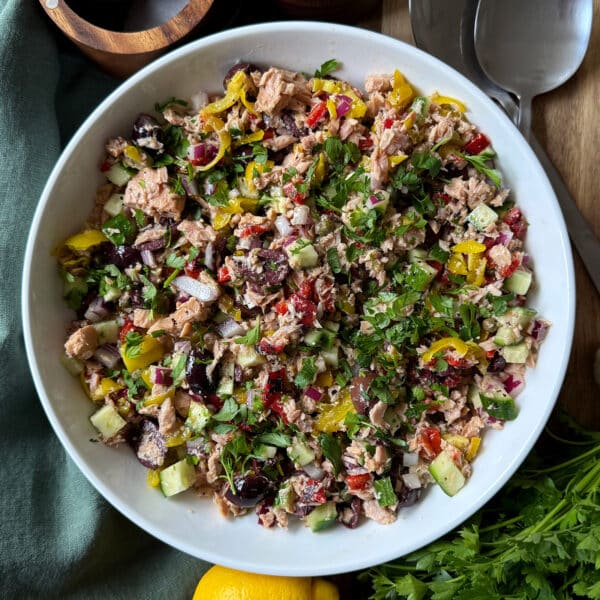 A white bowl filled with a colorful Mediterranean salad, including cucumber, olives, tuna, red onion, herbs, and pepperoncini. Fresh parsley, lemon, and serving utensils are visible nearby.