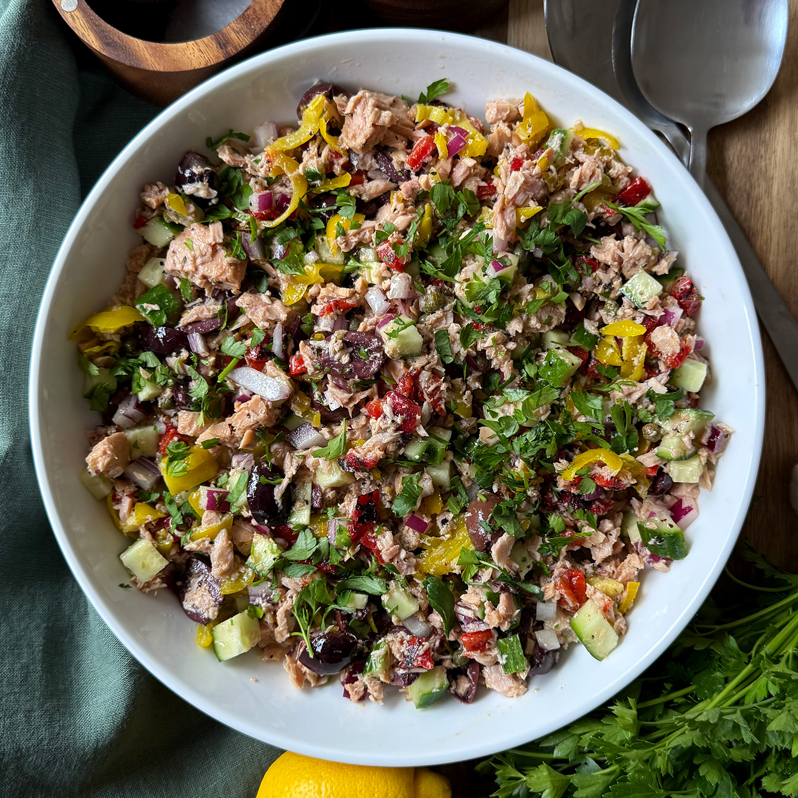 A white bowl filled with a colorful Mediterranean salad, including cucumber, olives, tuna, red onion, herbs, and pepperoncini. Fresh parsley, lemon, and serving utensils are visible nearby.