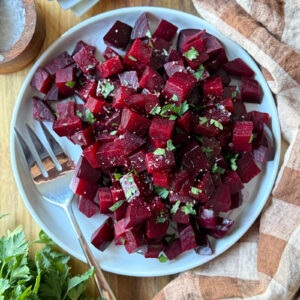 A white plate filled with cubed, roasted beets garnished with chopped herbs sits next to a fork. A striped napkin and fresh parsley are arranged nearby on a wooden surface.