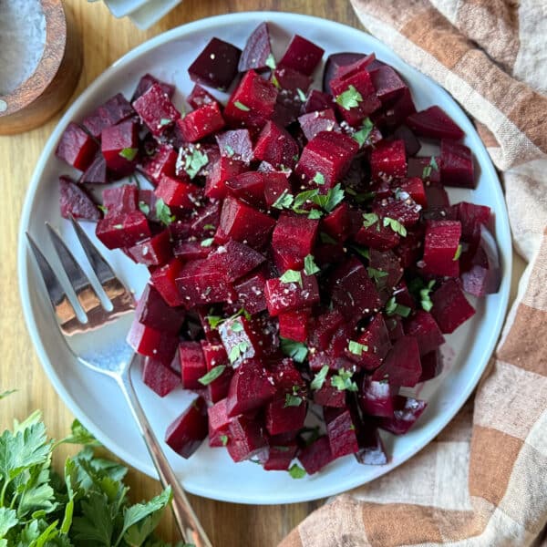 A white plate filled with cubed, roasted beets garnished with chopped herbs sits next to a fork. A striped napkin and fresh parsley are arranged nearby on a wooden surface.