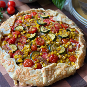 A rustic tomato galette with a golden crust, topped with zucchini, cherry tomatoes, corn, and cheese, sits on a wooden board.
