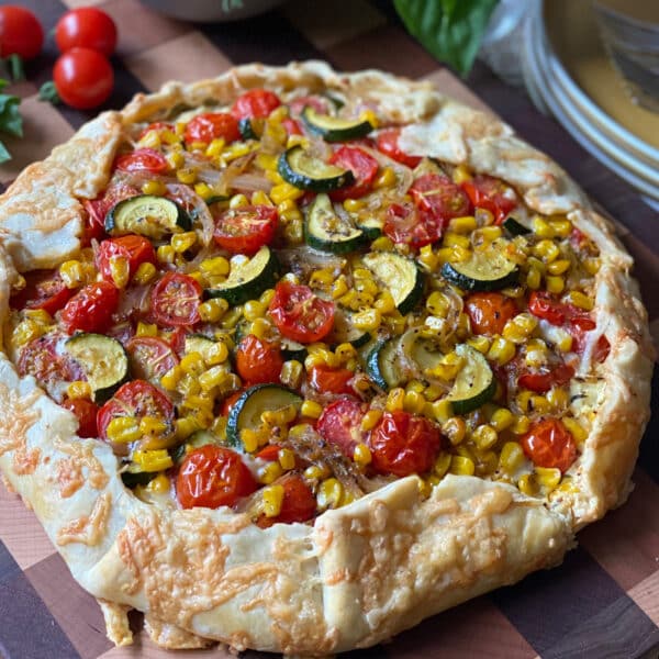 A rustic tomato galette with a golden crust, topped with zucchini, cherry tomatoes, corn, and cheese, sits on a wooden board.