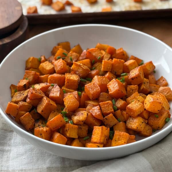 A white bowl filled with roasted, cubed sweet potatoes garnished with chopped chives, sitting on a light napkin with a wooden container and baking tray in the background.