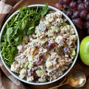 A bowl of tuna salad with grapes, celery, apples, walnuts, and arugula sits on a wooden tray, next to a bunch of red grapes, a green apple, and two gold spoons.