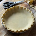 A homemade pie crust with crimped edges sits unbaked in a metal pie pan on a wooden surface. Kitchen scissors and scraps of dough are nearby, and a dusting of flour is visible on the board.