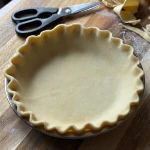 A homemade pie crust with crimped edges sits unbaked in a metal pie pan on a wooden surface. Kitchen scissors and scraps of dough are nearby, and a dusting of flour is visible on the board.