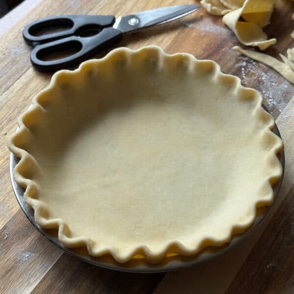 A homemade pie crust with crimped edges sits unbaked in a metal pie pan on a wooden surface. Kitchen scissors and scraps of dough are nearby, and a dusting of flour is visible on the board.