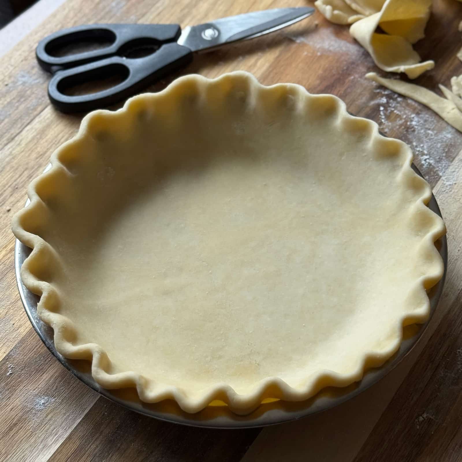 A homemade pie crust with crimped edges sits unbaked in a metal pie pan on a wooden surface. Kitchen scissors and scraps of dough are nearby, and a dusting of flour is visible on the board.