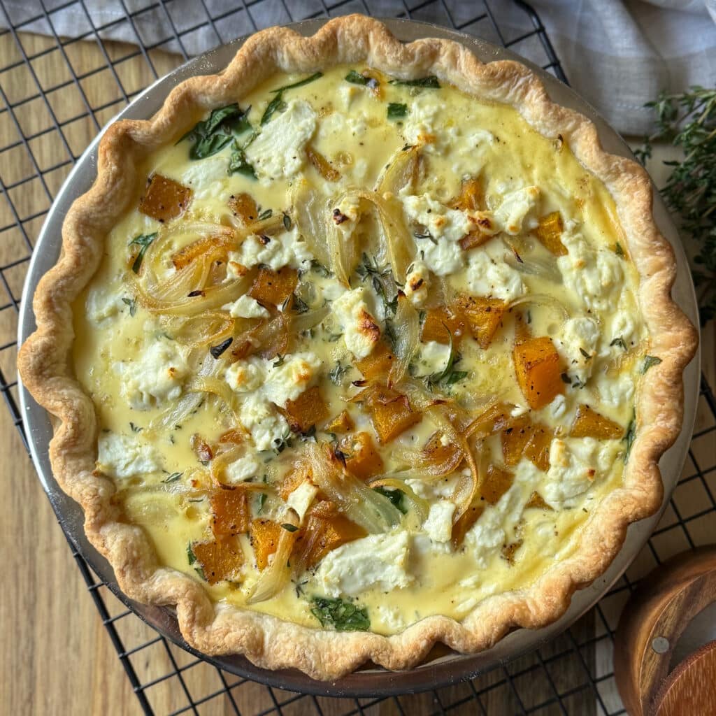A golden butternut squash quiche filled with roasted butternut squash, caramelized onions, spinach, and crumbled goat cheese, in a flaky crust, rests on a cooling rack.