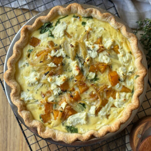 A golden butternut squash quiche filled with roasted butternut squash, caramelized onions, spinach, and crumbled goat cheese, in a flaky crust, rests on a cooling rack.