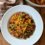 A bowl of chili cheese mac topped with shredded cheddar cheese and chopped parsley, placed on a wooden surface next to a brown plaid napkin.