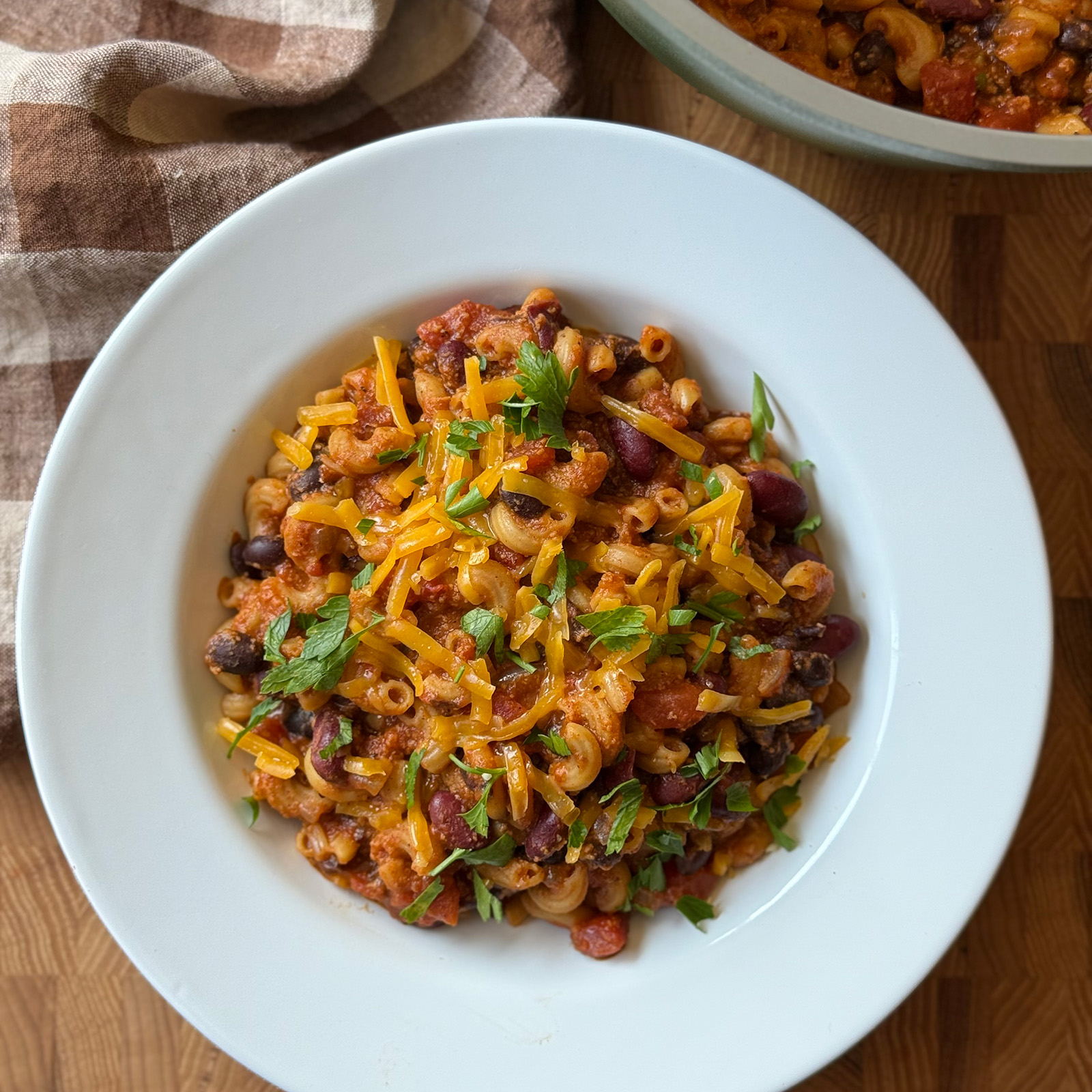 A bowl of chili cheese mac topped with shredded cheddar cheese and chopped parsley, placed on a wooden surface next to a brown plaid napkin.