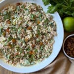 A bowl of curried tuna salad with herbs, raisins, and creamy dressing sits on a cloth. Fresh cilantro, a lime, and a small bowl of golden raisins are placed nearby.