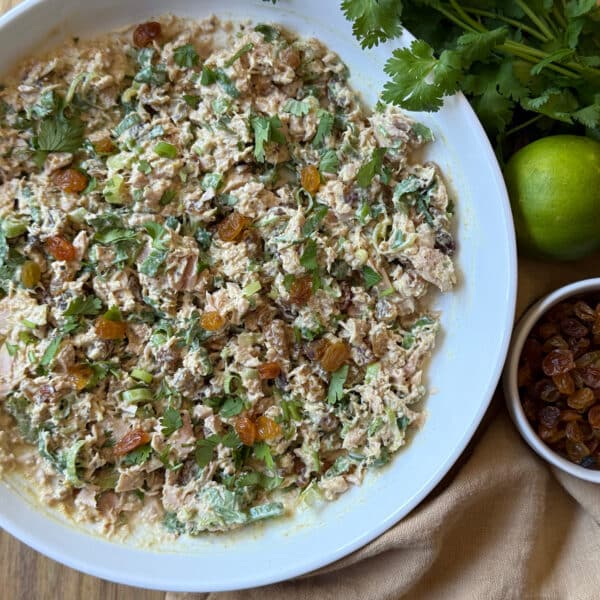 A bowl of curried tuna salad with herbs, raisins, and creamy dressing sits on a cloth. Fresh cilantro, a lime, and a small bowl of golden raisins are placed nearby.